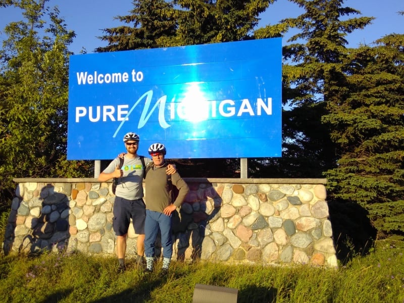 Chris and James are posing in front of a bib blue sign reading: Welcome to Pure Michigan