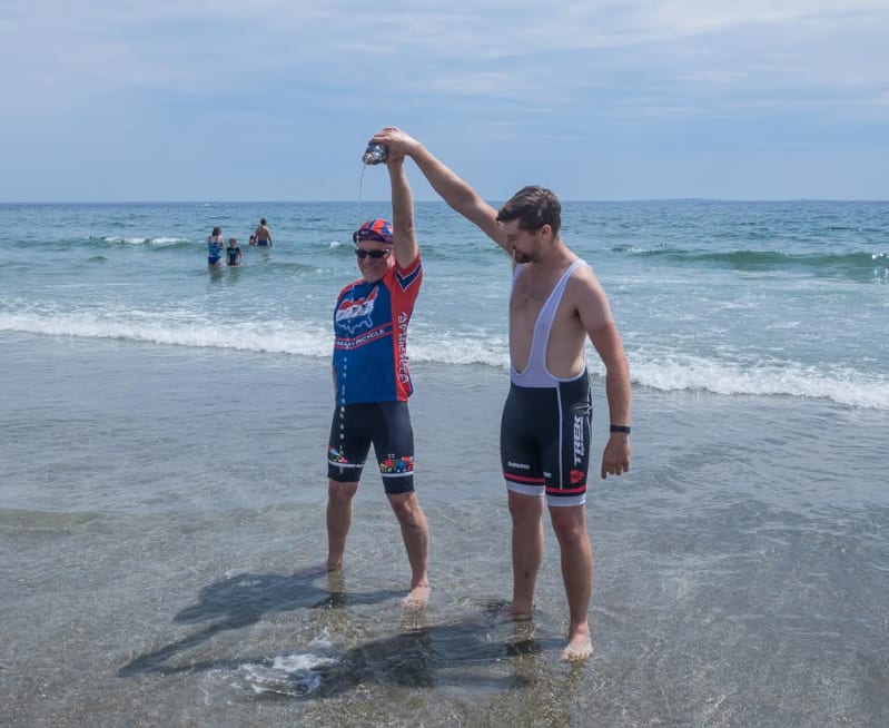 James and Chris are standing in the Atlantic up to their ankles, and raise a water bottle above their heads. Water is falling from the bottle. They look jubilant.